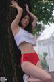 A woman in a white top and red shorts leaning against a tree.