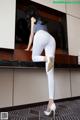 A woman in white leggings and high heels leaning against a counter.