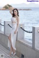 A woman in a white dress leaning against a wall by the water.